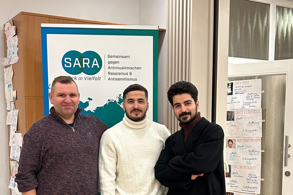 Drei Teilnehmer vor dem Aufsteller des Projekts SARA