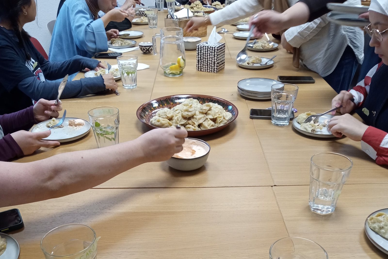 Lange Tafel mit Teilnehmerinnen beim Essen, in der Mitte eine Schale mit frisch gekochten Mantu, vorn ein Teller mit Mantu und Joghurtsoße
