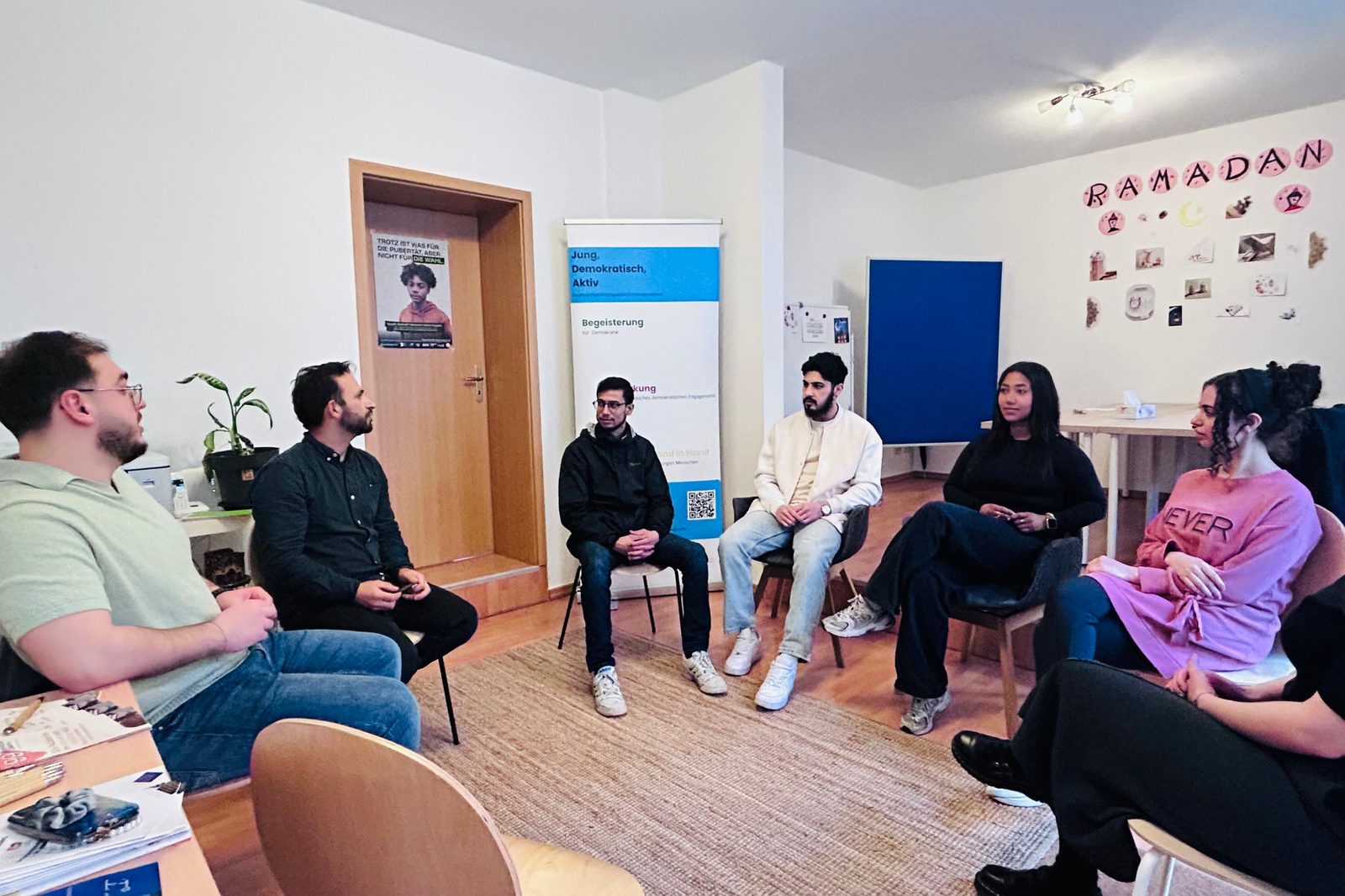 Junge Erwachsene im Stuhlkreis in den Vereinsräumen, an der Wand Ramadan-Bilder und der Aufsteller „Jung, Demokratisch, Aktiv“