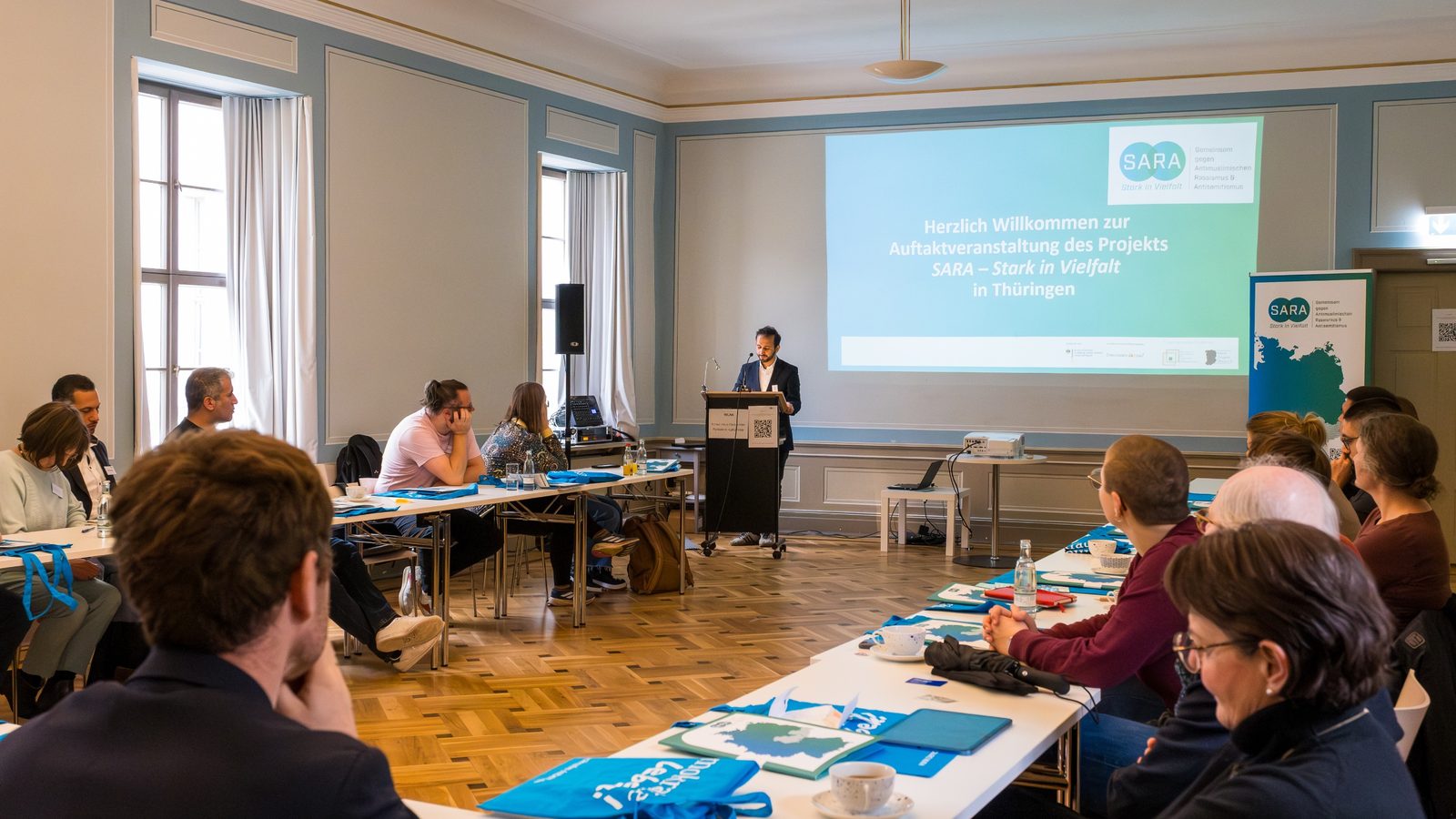 Gäste an langen Tischen im Saal, vorn am Rednerpult die Begrüßung vor der Folie „Auftaktveranstaltung des Projekts SARA – Stark in Vielfalt in Thüringen“